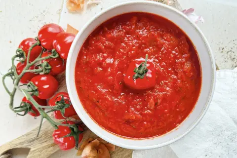 Eine Schüssel mit hausgemachter marinara sauce, garniert mit einer Kirschtomate, daneben frische Tomaten und Zwiebeln.