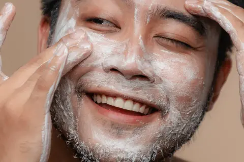 Un visage d'homme souriant, couvert de mousse nettoyante, se fait un soin du visage.