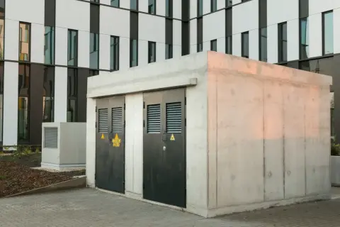 Betonowa stacja transformatorowa z dwoma czarnymi drzwiami i żółtymi znakami ostrzegawczymi. W tle nowoczesny budynek z białymi i ciemnymi panelami.