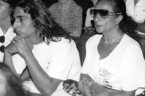 Un joven con pelo largo y una mujer con gafas de sol y joyas, sentados juntos. La imagen evoca la tristeza y el misterio en torno a como murio Lola Flores.