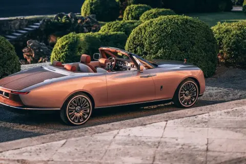 Elegancki Rolls-Royce Boat Tail w kolorze miedzianego różu, z drewnianym tyłem, zaparkowany wśród bujnej zieleni.