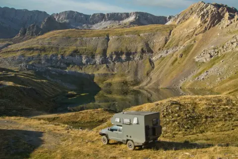 Una camper lista para la aventura en la Transpirenaica, aparcada junto a un lago de montaña con imponentes picos rocosos al fondo.