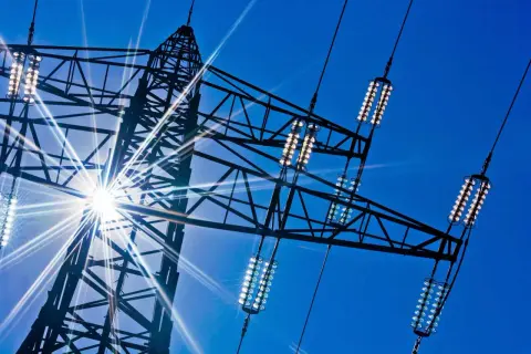 Słońce przebija się przez kratownicę wieży energetycznej, oświetlając izolatory przewodów, które przenoszą prąd dwufazowy.
