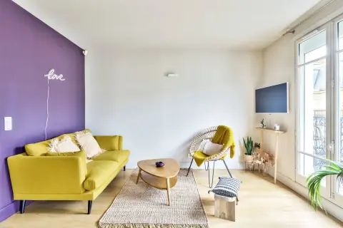 Ein gelbes Sofa und ein Korbsessel mit gelber Decke harmonieren perfekt mit der lila Wand. Welche Farbe passt zu lila? Hier sieht man, dass Gelb eine tolle Wahl ist!