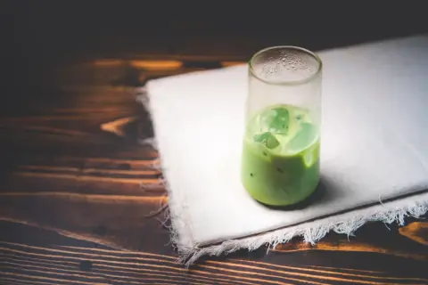 Un verre de matcha glacé sur un tissu blanc, sur fond de bois sombre. Une boisson rafraîchissante pour se demander : peut-on boire du matcha le soir ?