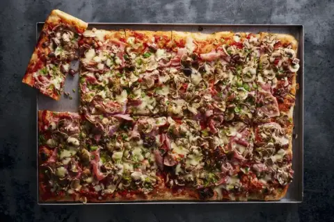Przepis na pizzę domową na blachę: prostokątna pizza z szynką, pieczarkami i oliwkami, posypana natką pietruszki.