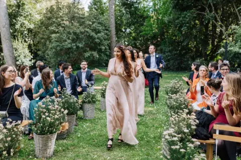 Le cortège d'église pour ce mariage en extérieur est joyeux. Les demoiselles d'honneur, en robes fluides, marchent sur l'herbe, applaudies par les invités.