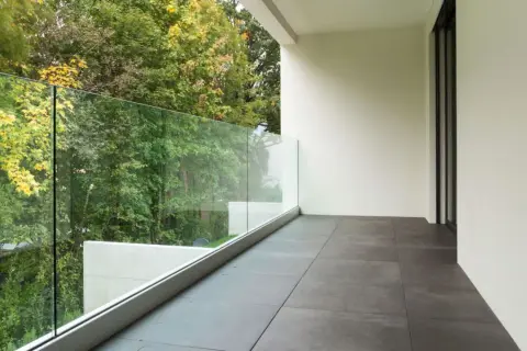 Nowoczesne płytki na balkon w bloku, z widokiem na zielony ogród i szklaną balustradę.