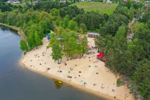 Plaża nad zalewem Cedzyna tętni życiem. Ludzie odpoczywają na piasku, ciesząc się słońcem i widokiem na jezioro.