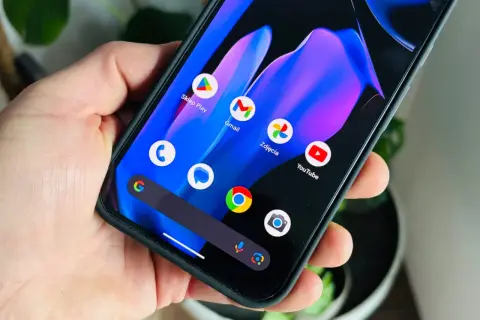 Dłoń trzyma smartfon z kolorową tapetą i ikonami aplikacji, w tym Sklep Play, Gmail, Zdjęcia i YouTube. Wygląda jak nowy android 15.