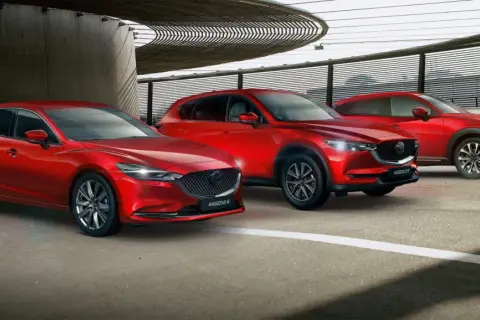 Trzy czerwone samochody marki Mazda: sedan, SUV i crossover, zaparkowane pod nowoczesną konstrukcją.