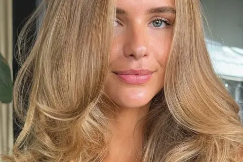 Piękna kobieta z falowanymi włosami w odcieniu maślany blond. Jej oczy są zielone, a usta delikatnie umalowane.