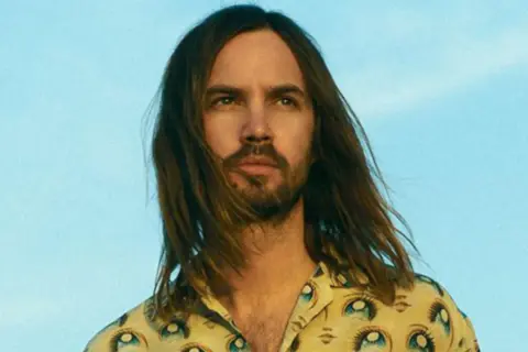 Kevin Parker w koszuli z motywem oczu, z rozwianymi włosami na tle błękitnego nieba.