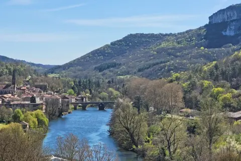 Vue panoramique de la gorge de l'Aveyron avec un village pittoresque, une rivière bleue et des collines verdoyantes sous un ciel clair.