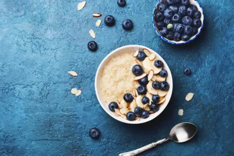 Porridge ricetta: una ciotola di avena con mirtilli, banane e mandorle a lamelle su sfondo blu.