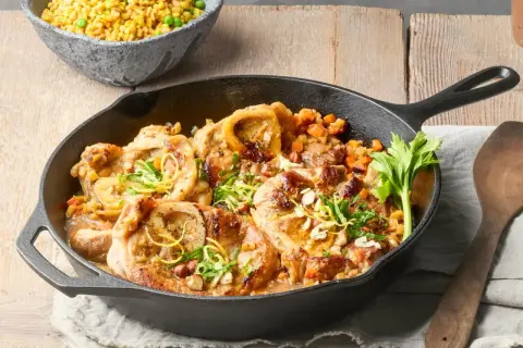 Herzhaftes Ossobuco alla Milanese in Gusseisenpfanne, garniert mit Zitronenzeste und Kräutern, daneben eine Schüssel Reis mit Erbsen.