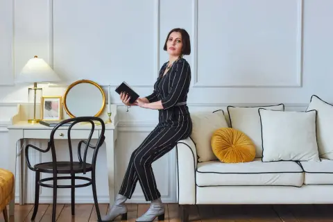 Elegancka kobieta w paski, w stylu glamour ubrania, siedzi na sofie, trzymając książkę. Obok biurko z lampą i lustrem.