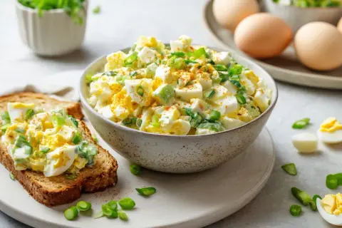 Cremiger Eiersalat mit Joghurt, garniert mit Frühlingszwiebeln, auf einer Scheibe Toastbrot. Daneben eine Schüssel mit dem Salat und gekochte Eier.