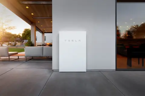 Un elegante Tesla Powerwall blanco se alza junto a una casa moderna con un patio y muebles de exterior.