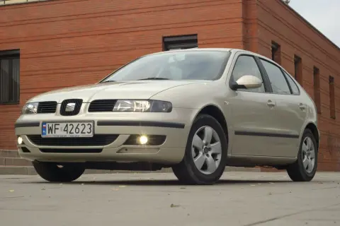 Jasny Seat Leon 1 z włączonymi światłami mijania i przeciwmgielnymi, zaparkowany przed ceglanym budynkiem.
