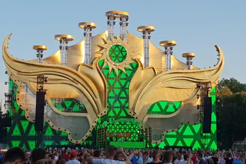 Złota, słoneczna scena festiwalowa z zielonymi światłami, gdzie jest sunrise, otoczona tłumem ludzi.
