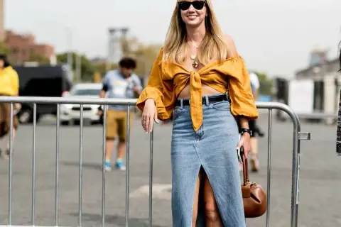 Mujer sonriente con un outfit falda vaquera larga, blusa amarilla anudada y bolso marrón.