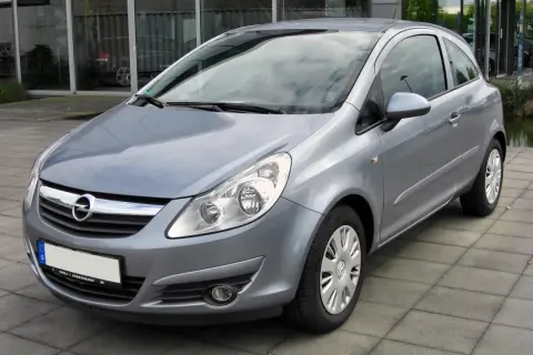 Srebrny Opel Corsa D 1.4 benzyna stoi na chodniku przed budynkiem z dużymi oknami.