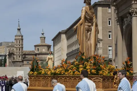 Procesión de **domingo de resurrección Zaragoza**. Figuras religiosas adornadas con flores en un paso, ante edificios históricos y una multitud.