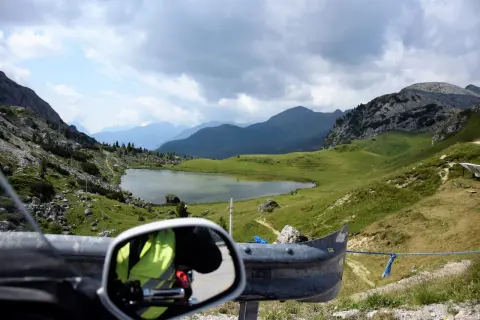 Widok z motocykla na malownicze **przełęcze alpejskie**, jezioro i góry pod zachmurzonym niebem.
