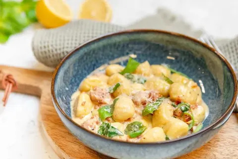 En skål med krämig gnocchi salsiccia, toppad med färsk basilika och parmesan. Citroner och en grå handduk i bakgrunden.