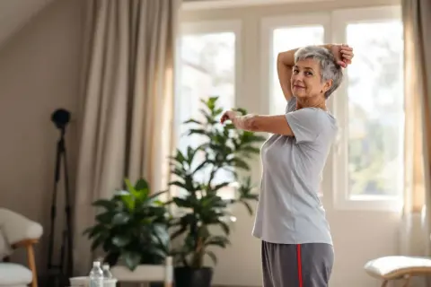 Une femme senior souriante pratique un assouplissement du dos, bras levés, dans un salon lumineux.