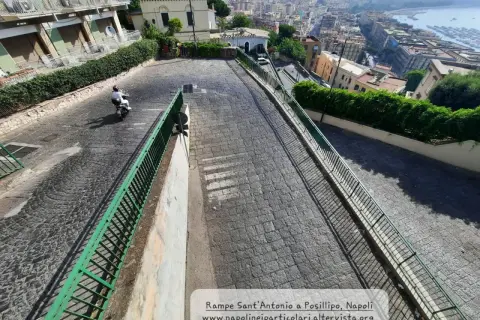 Le 13 discese di Napoli, una vista panoramica di una strada in discesa con un motorino, tra palazzi e vegetazione, che porta verso il mare.