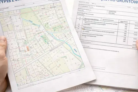 Ręce trzymają wypis i wyrys z rejestru gruntów. Obok leży kalkulator. Dokumenty zawierają mapę działek i tabelę z danymi.