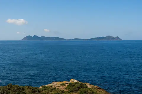 El mar azul se extiende hasta las islas de **cabo Estai**, con una costa rocosa y vegetación en primer plano.