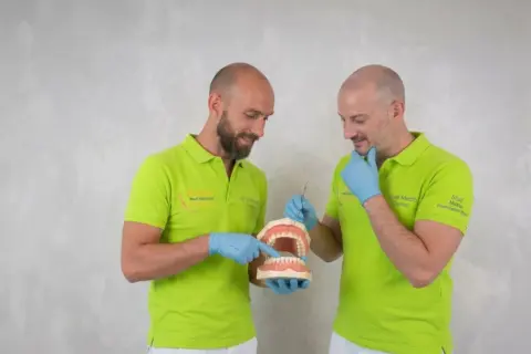 Due dentisti con camici verdi mostrano un modello di denti, spiegando come si chiamano i denti davanti.
