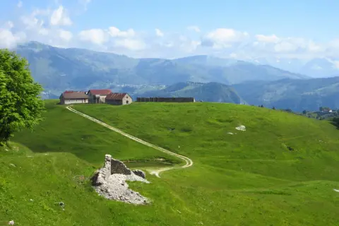 Un sentiero delle malghe serpeggia tra verdi colline, conducendo a un gruppo di rustici edifici con tetti rossi. In primo piano, ruderi di pietra e un piccolo stagno.