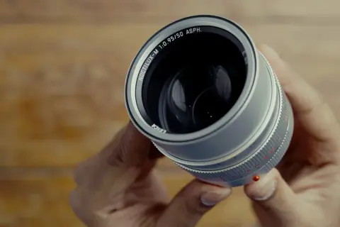 Hands hold a Leica Noctilux M 50 mm lens, its silver barrel and prominent f/0.95 aperture gleaming.