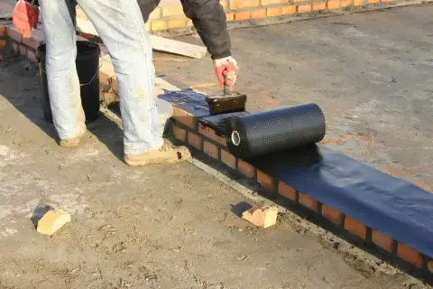 Pracownik kładzie hydroizolację na murze, przygotowując zbrojenie pod dalsze prace budowlane.