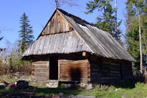 Drewniana bacówka w górach, otoczona drzewami, pod błękitnym niebem.