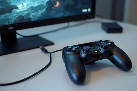 Czarny pad PS4 podłączony kablem do komputera, obok monitor z grą.