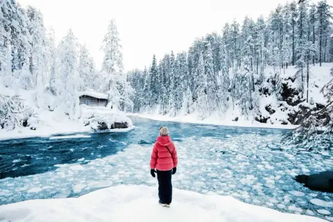 Une personne en manteau rouge admire un paysage hivernal de la pays Lapone, avec une rivière gelée et une cabane enneigée.