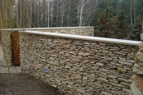 Solidne ogrodzenie z piaskowca, z drewnianymi drzwiami i kamiennymi lampkami, otacza ogród.