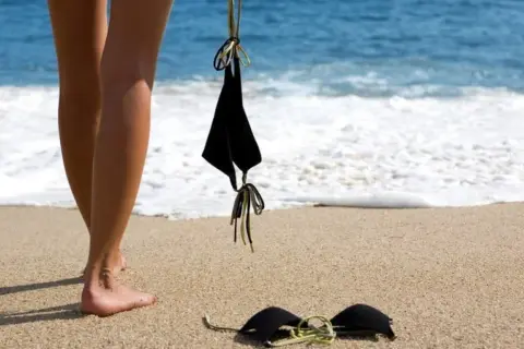 Nogi na piaszczystej plaży, obok czarny biustonosz. W tle błękitne morze i biała piana fal. Idealne miejsce na plażę nudystów.