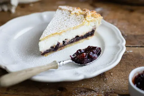 Una fetta di crostata ricotta e visciole, cosparsa di zucchero a velo, su un piatto bianco. Un cucchiaino è pieno di visciole sciroppate.