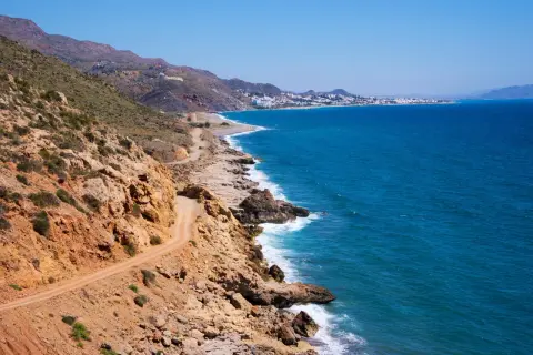 Costa rocosa con camino de tierra y el mar azul. Al fondo, se divisa la silueta de Mojácar y sus playas.