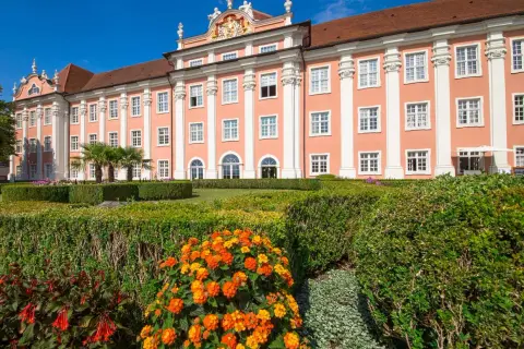 Das Meersburg Neues Schloss erstrahlt in Rosa vor blauem Himmel. Ein gepflegter Garten mit bunten Blumen und Palmen lädt zum Verweilen ein.