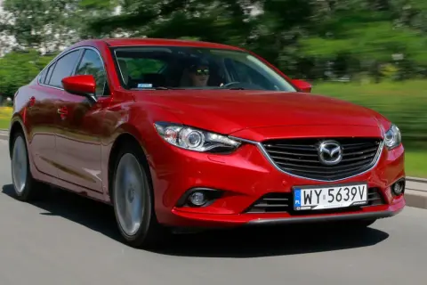 Czerwona Mazda 6 w ruchu ulicznym. Kierowca w okularach przeciwsłonecznych. Wiele pozytywnych mazda 6 opinie potwierdza jej sportowy charakter.
