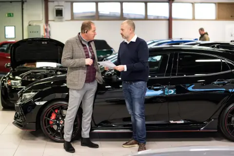Två män diskuterar papper vid en svart sportbil. De verkar prata om bilkostnad per månad.
