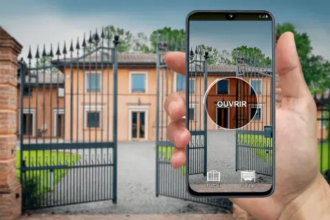 Une main tient un iPhone affichant une application de télécommande universelle pour ouvrir le portail d'une villa.