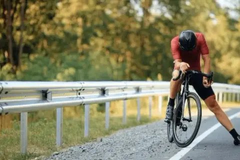 Cycliste en tenue de sport, casque noir, s'appuyant sur son vélo sur le bord de la route. Il semble fatigué, peut-être après avoir fait du sport à jeun.